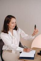 The aromatherapist girl is sitting in her office and holding a bottle of aromatic oil in her hands and writing something down. there are essential oils on the table photo