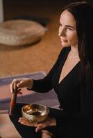 A woman in the lotus position using a singing bowl indoors . Relaxation and meditation. Sound therapy, alternative medicine. Buddhist healing practices photo