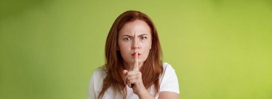 Not speak during exam. Strict serious-looking displeased middle-aged redhead woman frowning disappointed hushing say shush index finger pressed lips keep quiet gesture green background photo