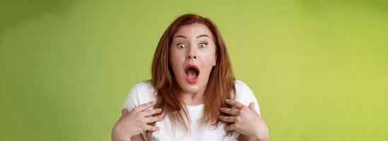 Shocked panicking redhead middle-aged woman gasping drop jaw open mouth stare camera freak-out anxious pointing herself impressed terrified frustrated nervously react green background photo