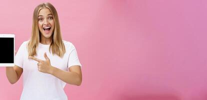 Woman giving recommendation buy digital tablet pointing at device screen with index finger smiling enthusiastic and pleased telling own opinion about gadget perfomance posing delighted over pink wall photo