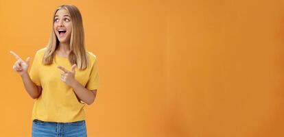 Amused enthusiastic and charismatic girl with blond hair and tanned pure skin without makeup pointing and looking at upper left corner with joyful happy smile over orange background photo