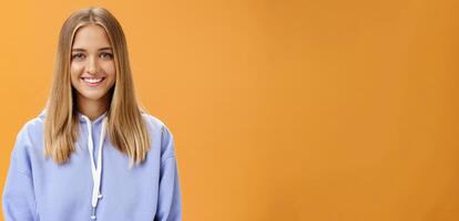 Waist-up shot of pleasant kind and happy friendly-looking young fair-haired girl in comfortable hoodie smiling cheerfully at camera with delighted carefree look posing against orange background photo