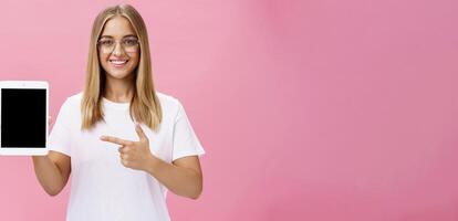 Waist-up shot of optimistic and joyful female showing cool digital tablet pointing at device screen and smiling broadly at camera giving advice what app useful posing in glasses against pink wall photo