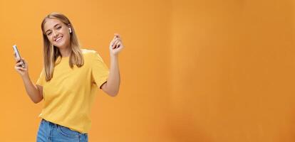 Joyful carefree european woman wearing wireless earbuds holding smartphone dancing with raised hands and pleased happy smile leading vanlife having time to relax and enjoy life over orange wall photo