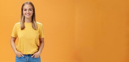 Cute chubby female with tanned skin and fair hair posing optimistic and joyful against orange background holding hands in pockets smiling broadly at camera charismatic and friendly-looking photo