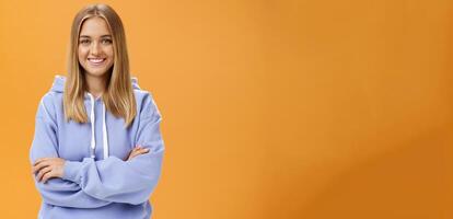 Young creative and ambitious female student staring own project at home working freelance having free time and working when want holding hand crossed on chest wearing cozy hoodie over orange wall photo