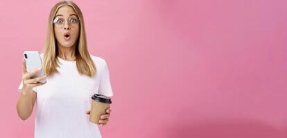 Girl receiving awesome message on smartphone while drinking coffee from cup in cafe sharing news with friend folding lips in wow sound staring surprised at camera holding cellphone over pink wall photo