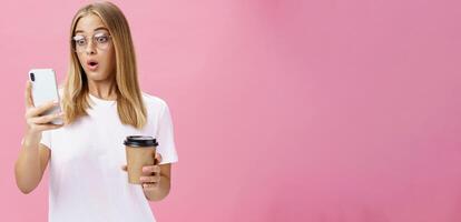 Woman drinking coffee being shocked by received message reacting on stunning news folding lips gasping looking astonished and impressed at smartphone screen holding paper cup, posing over pink wall photo