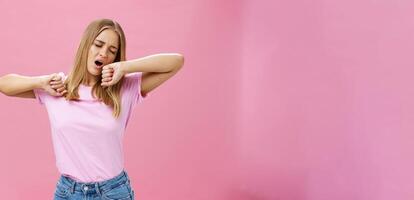 Too tired to work today. Lazy and exhausted attractive young female student doing homework all night yawning with closed eyes while stretching hands from after tiresome project over pink wall photo