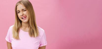 Playful flirty attractive young girl with fair straight hair in t-shirt winking at camera smiling and licking upper lip with tongue feeling enthusiastic and carefree against pink background photo