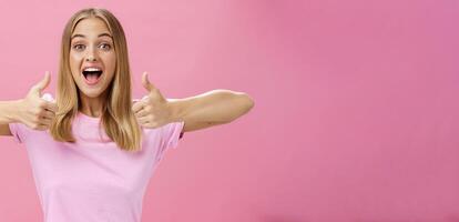 Supportive girlfriend cheering for favorite player showing thumbs up with happy positive smile giving approval and showing she likes perfect idea posing amused and enthusiastic against pink background photo