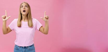 Portrait of amused and impressed curious attractive woman in t-shirt dropping jaw looking and pointing up astonished and intrigued watching interesting object in sky posing against pink background photo