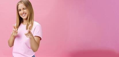 Woman expressing positive attitude towards camera pointing with fingers and winking joyfully smiling being uplifted, standing in good mood with optimistic gestures against pink background photo