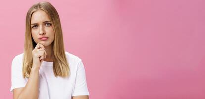 Girl facing problem thinking being hesitant while making decision holding hand on chin frowning pursing lips standing thoughtful over pink background in white casual t-shirt photo