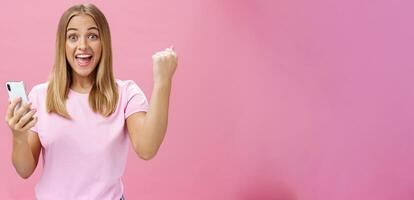 Girl beat own record in smartphone game raising clenched fist in cheer and triumph holding cellphone, smiling excited and happy at camera celebrating victory with joyful gesture over pink background photo