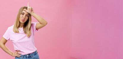 Girl mocking under boyfriend showing loser sign on forehead sticking out tongue gazing arrogant and indifferent at camera being moody holding hand on waist posing against pink background photo