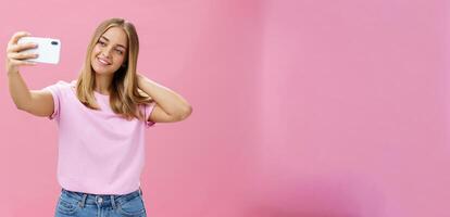 Female beauty blogger taking selfie post new look online. Portrait of charming tanned young woman in t-shirt touching hair gently pulling hand with smartphone near face taking photo over pink wall
