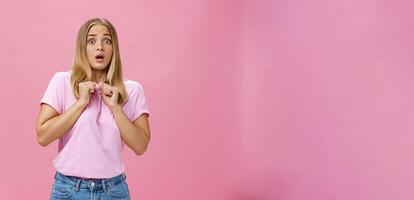 Woman gasping from fright being scared by someone pressing clenched hands to chest bending backwards looking worried and terrified at camera being shocked with unexpected bang over pink wall photo