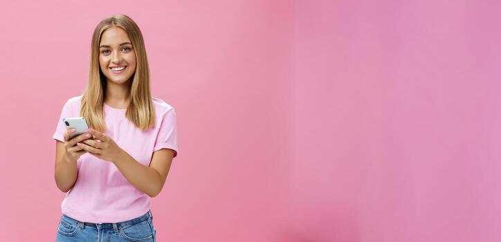Woman calling taxy via smartphone asking friend address smiling cheerfully at camera holding phone with both hands over chest getting in touch with clients via messages standing over pink wall photo