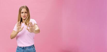 Stay away from me. Intense worried and displeased woman being victim of sexual harassment raising palms in defensive gesture making step backwards frowning asking stop, refusing over pink wall photo