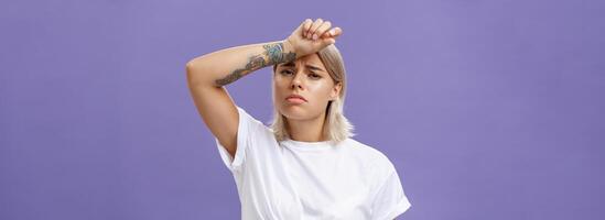 Indoor shot of tired and displeased exhausted cute european woman with tanned skin and tattoos on arms holding hand on forehead frowning suffering from tiresome work or sun heating over purple wall photo