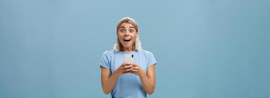 Indoor shot of amazed attractive blonde female coworker in casual t-shirt gasping from amazement and happiness being surprised staring surprised at camera holding smartphone receiving awesome news photo