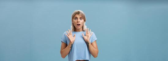 Shocked and worried concerned good-looking feminine woman with blond hair and tattooed arms raising palms gasping and opening mouth expressing empathy and feeling sorry for friend over blue wall photo