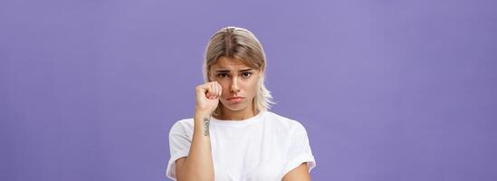 Studio shot of offended sad and timid silly woman with blond hairstyle frowning looking from under forehead holding fist near eye as if whiping teardrop being upset over purple background photo