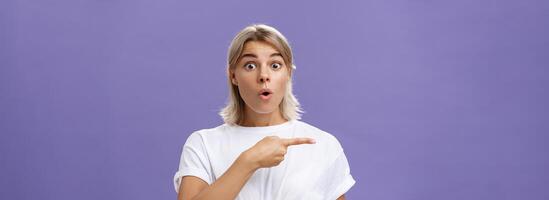 Waist-up shot of overwhelmed excited and surprised attractive urban female in white t-shirt folding lips gasping from amazement being questioned asking question and pointing left over purple wall photo