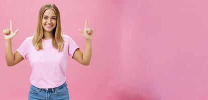 Indoor shot of pleasant attractive friendly-looking girl with tanned skin in casual t-shirt and jeans raising hands pointing up and smiling broadly at camera with satisfied look over pink background photo