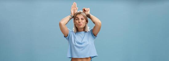 Flirty attractive and stylish cool girlfriend with tattooed arms and pierced belly winking folding lips in air kiss showing bunny ears with palms on head standing over blue background playful photo