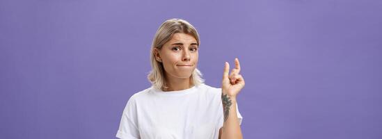 Girl having tiny problem. Concerned attractive blonde girl with tattoo on arm pursing lips in troubled look shaping small or little object, dissatisfied with regret in eyes over purple wall photo