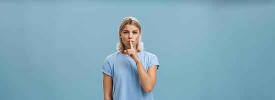 Promise not tell anyone it secret. Mysterious attractive and feminine blond woman with tanned skin and tattoos shushing with index finger over mouth squinting as if hiding something over blue wall photo