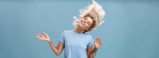 Waist-up shot of artistic and creative attractive carefree blonde female waving head and hair joyfully dancing and jumping having fun smiling broadly over blue background photo