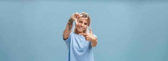 Be creative and happy. Portrait of charming enthusiastic female designer with blond hair and tattoo on arm winking and smiling joyfully making frame gesture as if measuring space over blue background photo