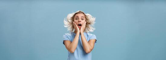 Indoor shot of shocked and impressed young stylish european female with blond hair holding hands on cheek from empathy and surprise opening mouth facing wind posing with hairstyle floating in air photo