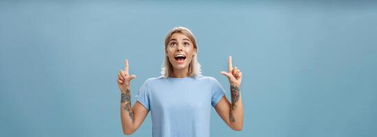 Studio shot of impressed speechless happy good-looking woman with tattoos on arms dropping jaw from amazement and joy gazing fascinated and pointing up standing over blue background photo