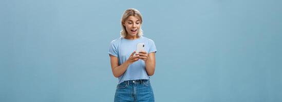 Entertained charming young european female with blond hair in casual outfit holding smartphone being satisfied with great camera and photos gasping and gazing amazed at screen over blue wall