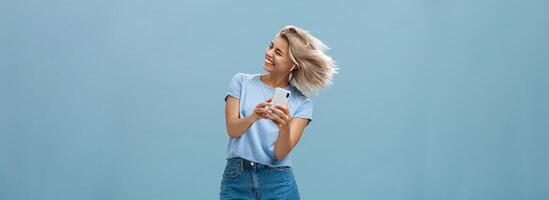 Great vibes carrying me away. Carefree joyful attractive young blonde female in stylish outfit waving head and smiling broadly listening music in wireless earbuds and holding smartphone over blue wall photo