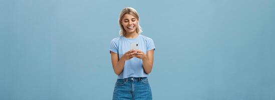 Nice to have internet friends. Carefree happy attractive blonde female student with tanned skin and blond hair reading message from smartphone smiling at device screen pleased over blue wall photo