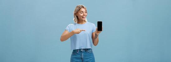 Portrait of delighted good-looking european female with blond haircut showing smartphone screen with amazement and delight pointing at device while promoting brand new gadget over blue background photo