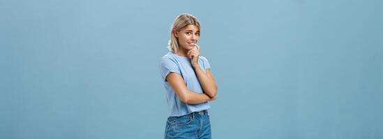 Studio shot of unconfident silly and insecure cute feminine girl with blond hair standing half-turned over blue background holding finger on lip while being shy ask question smiling hesitating photo