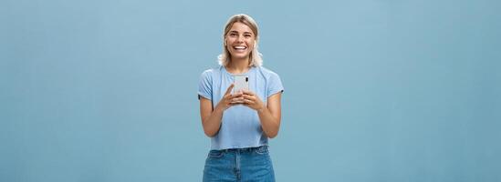 Satisfied happy and carefree young attractive blonde woman in casual outfit smiling broadly from happiness and delight listening music in wireless earphones holding smartphone over blue wall photo