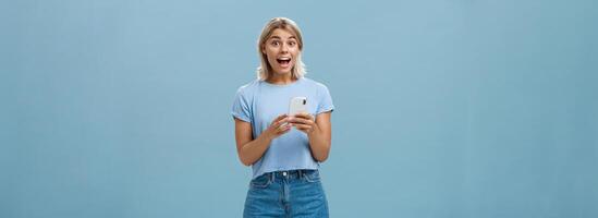 So happy receive premium for free reacting with enthusiasm and excitement on amazing message smiling broadly with delight and surprise holding smartphone posing over blue background photo