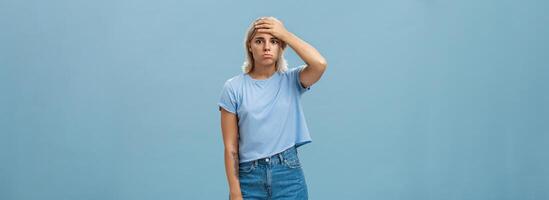 Concerned and troubled young teenage girl being shocked with loads of responsibilities holding hand on forehead staring spaced out and pouting from tiresome perplexed situation over blue wall photo