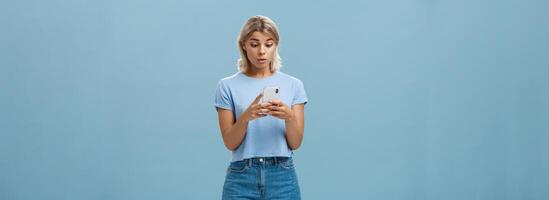 Indoor shot of surprised young caucasian girl receiving unexpected invitation via smartphone reading strange message in cellphone staring stunned and confused at screen over blue background photo