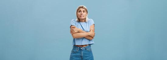 Turn off air-cooler I am freezing in summer. Displeased cute caucasian blonde girl in trendy t-shirt hugging herself while trembling from cold shaking gazing right with worried look over blue wall photo