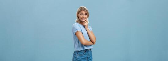 Indoor shot of feminine romantic and timid good-looking european female with fair hair holding finger on lip smiling curious and intrigued while turning right and observing interesting thing photo