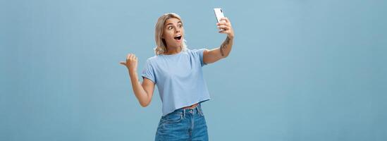 Girl recording video showing followers awesome scene attending cool event holding smartphone looking impressed and surprised at device screen pointing backwards indicating right over blue wall photo